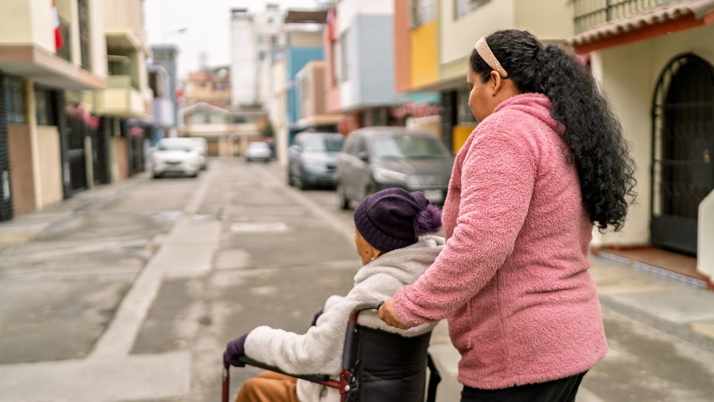 Mujer acompaña a una adulta mayor en silla de ruedas por una calle colorida de una ciudad latinoamericana